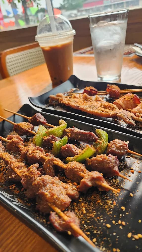 Lamb and green-pepper skewers piled on a black plate next to an iced milk tea.