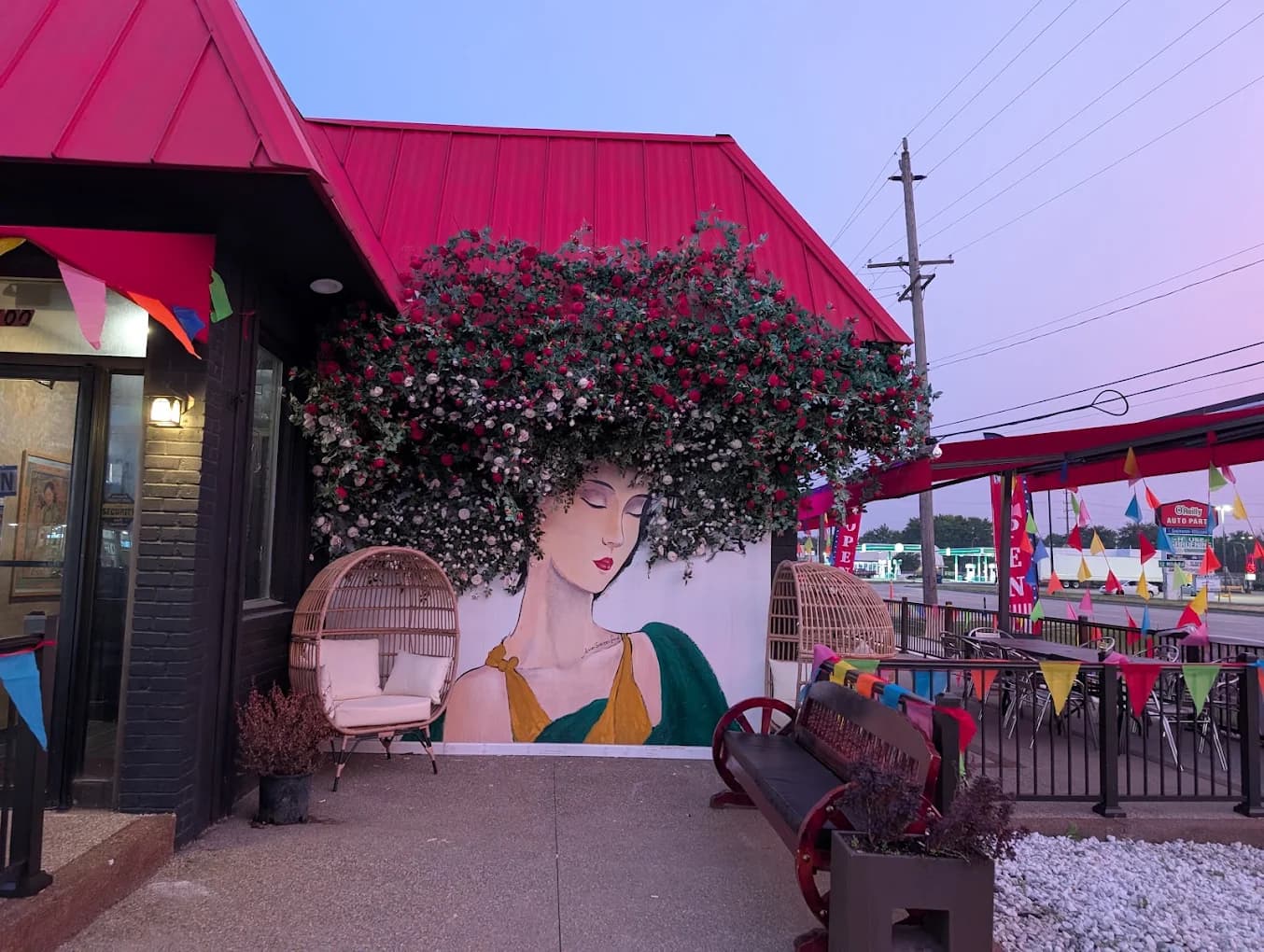 ASF patio at dusk: a mural of a woman whose hair is a bloom of red and white roses, framed by a red roof, egg chairs and colorful bunting.
